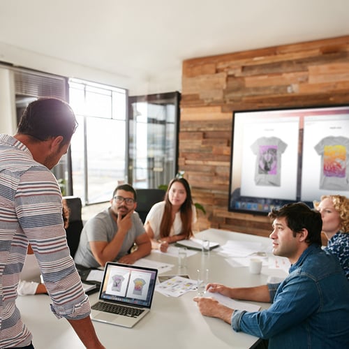 A team of people meeting in a room. One person is wirelessly mirroring their computer screen to a larger display.